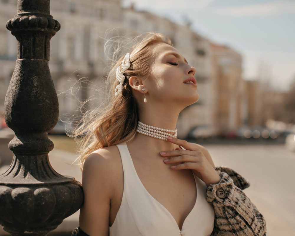 Portrait of beautiful blonde lady in stylish white silk dress, checkered jacket and pearl necklace Portrait of beautiful blonde lady in stylish white silk dress, checkered jacket and pearl necklace gently touching neck and posing at city square.
